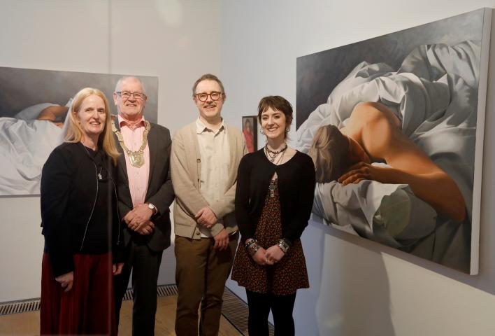 4 people are standing in a line next to a painting. From left to right there is a woman with shoulder length light red hair, then a man with short grey hair and he is wearing an official gold chain around his neck that comes down to the middle of his chest, then another man with glasses and a young woman with short brown hair and lots of necklaces. All are smiling at the camera.