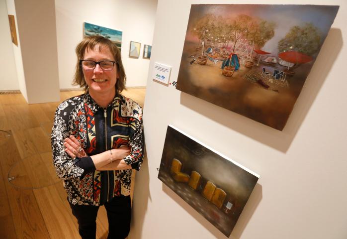 Artist Claire Halpin has short brown hair and glasses. She is standing with her arms crossed next to her two paintings which are on the right. The top painting shows a landscape with tents and trees but is empty of people. The lower painting shows a line or 5 yellow portaloos. one is knocked over and there is rubbish on the floor and hints of bullet holes in the yellow doors of the toilets.