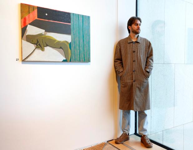 Photo of artist Fergal Styles next to his painting of a woman asleep on a bed. Fergal has brown hair and is wearing a ling light brown trenchoat and is standing to the right of the painting in a corner, one side of the corner is a glass wall. He is looking out the glass wall to the right.