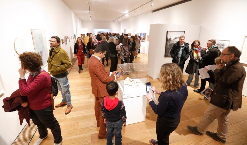A gallery with white walls is filled with people looking at the art and talking in small groups.