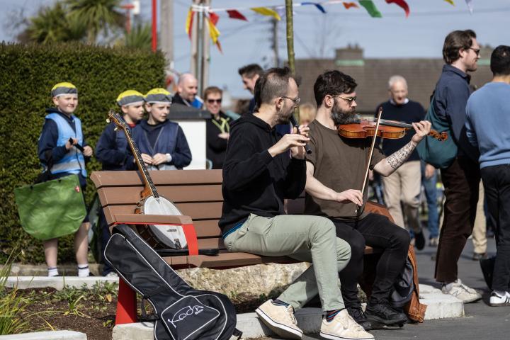 2 men sitting on a bench playing violin &amp; flute surrounded by group of adults and kids