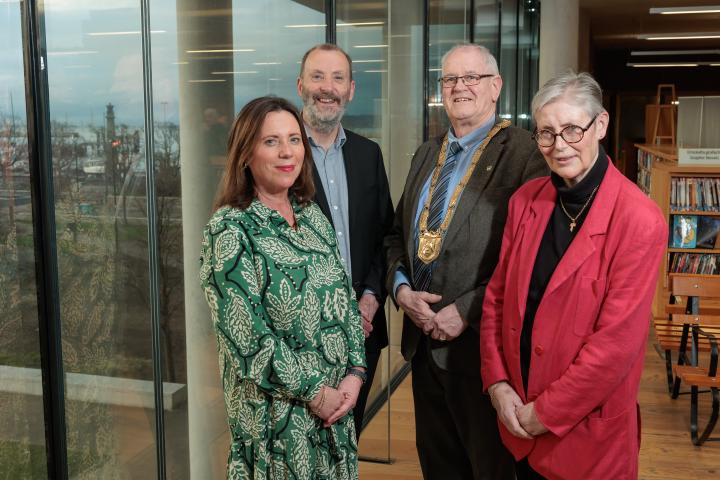Four people standing indoors at an event space, beside large windows with shelving and books in the background.