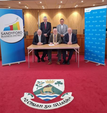 Five representatives from Dún Laoghaire–Rathdown County Council, Sandyford Business District and NovaUCD seated and standing at a table during the signing of a Memorandum of Understanding