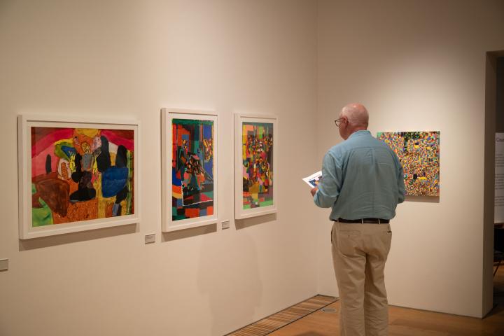 An older white man in a light blue short and khaki pants has his back to the camera as he views 3 colourful paintings on a white wall in a gallery space.