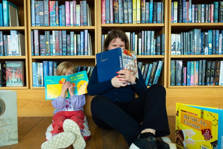 A picture of a woman and a small child sitting at the library covering their faces with books. The background is bookshelves.