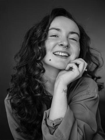 black and white photo of a young white woman with ling curly dark hair. she is smiling at the camera and has her chin resting in her hand.