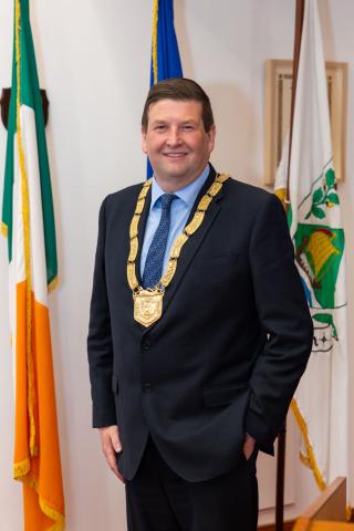 man standing in front of Irish flag wearing chain of office
