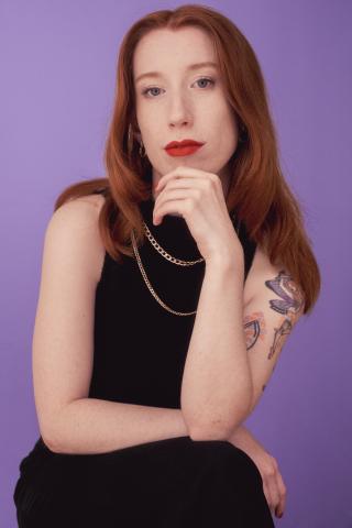 colour photo of a female presenting person looking directly at the camera. They have their legs crossed and an arm resting on their knee supporting their chin. They have shoulder length red hair, bright red lipstick. they are wearing a black sleeveless top which shows tattoos on one shoulder.