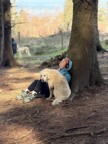 a woman sits on the ground under a tree with her head resting on the tree. it's in a forest and more trees are visible in background. A golden retriever dog sits beside her. she is listening to something on headphones