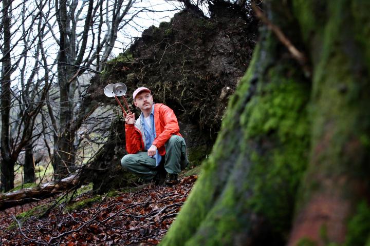 A man in a bright red/orange raincoat with a cap on his head is crouched in a forest setting at the base of a tree