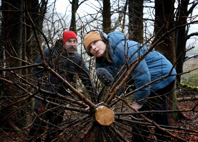 a woman in a blue jacket and yellow hat is wearing headphones and is bent over looking upwards through some branches. A man in a red hat is behind her and is also looking up. There is a microphone positioned on the branches, they are in a forest