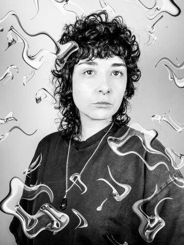 Black and white photo of a white female presenting person with short curly dark hair. They are wearing a dark top with a necklace hanging low on their chest. Over the photo is a digital effect that looks like water waves.