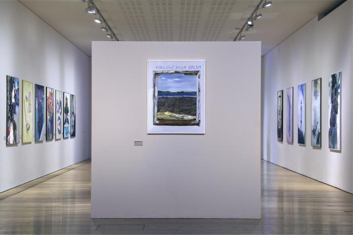 photo of an art exhibition. In the foreground is a freestanding white wall with a painting displayed on it. Behind the wall is a room with white walls and wooden floor. either side of the room has paintings on the walls.