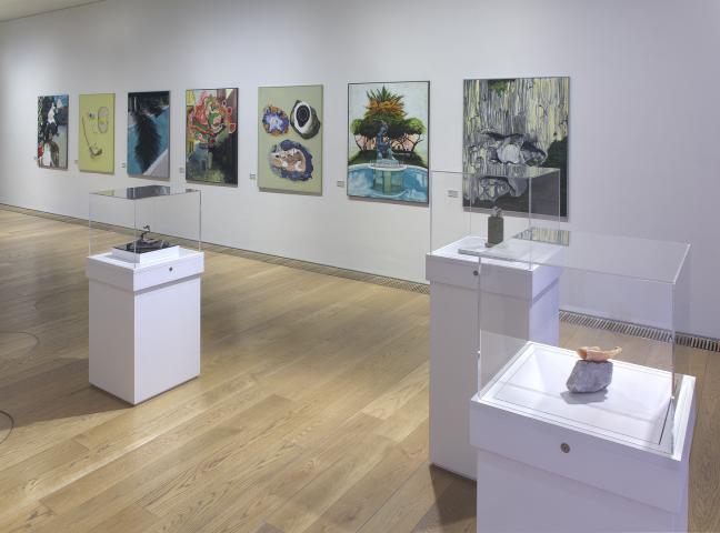Photo of an art exhibition. A long white wall with a row of paintings is at the back of the room. The floor is a light coloured wood. In the middle of the room are three white plinths with clear Perspex boxed on top. In the boxes are small sculptures.