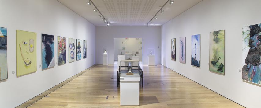 photo of an art exhibition. The room is long with white walls and wooden floor. either side of the room has paintings on the walls.