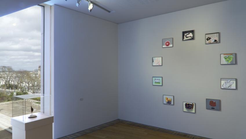 Photo of an art exhibition. It shows the corner of a room with white walls. the wall on the left has a floor to ceiling window and an empty wall. The wall on the right has 10 small paintings arranged in a circle. To the left is a small plinth with a small sculpture displayed in it.