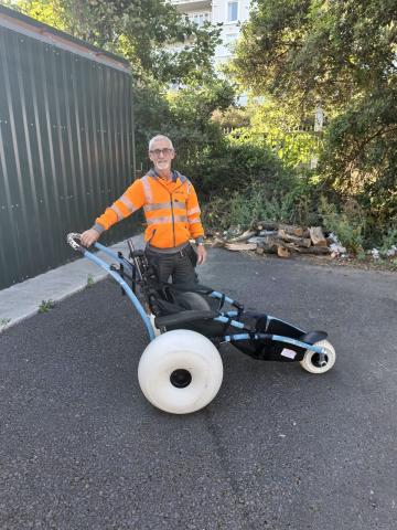 A man standing with his right hand on the top of a hippocampe beach wheelchair with enlarged wheel to allow for all terrain use
