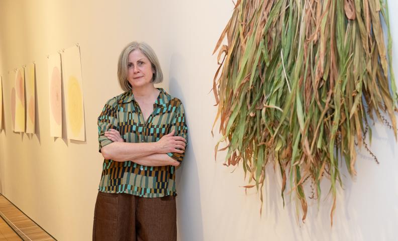 An individual stands with folded arms beside a wall-mounted installation of dried foliage in an art gallery. A row of abstract circular artworks is displayed along the wall in the background.