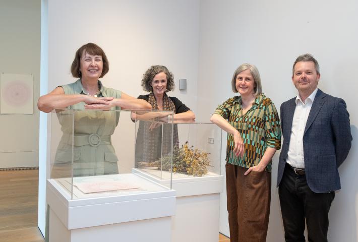 Three women and a man stand beside glass display cases in a contemporary art gallery. The cases contain artworks, including dried plant material and paper pieces, with additional artwork displayed on the walls behind them.