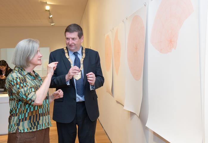 Two people converse while viewing a series of large abstract circular artworks hanging on a gallery wall. One person wears a ceremonial chain, and the exhibition space is lit by overhead gallery lighting.