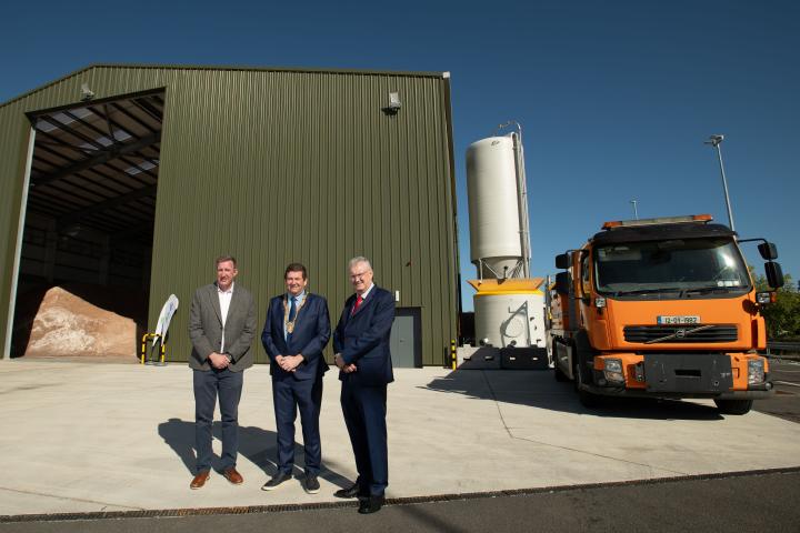 Group of dlr staff standing outside an industrial facility beside an orange gritting truck and a large brine storage tank during a site visit on a sunny day