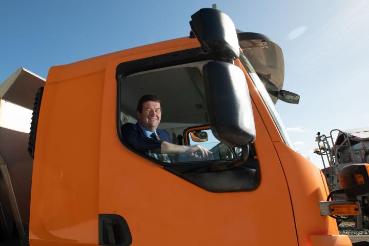 Councillor Barry Saul seated in the cab of an orange gritting truck, viewed through the driver’s side window, with a clear blue sky and depot machinery in the background.
