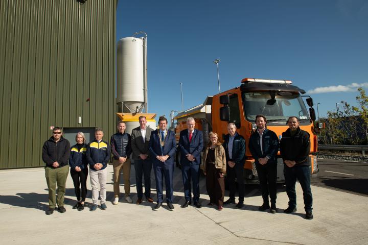Group photograph of staff and stakeholders standing outside the Ballyogan brine manufacturing facility beside an orange gritting truck and a large brine storage silo.