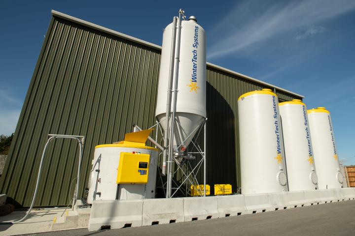Brine manufacturing equipment including a large storage silo, four white storage tanks with yellow tops, and associated pipework beside a green industrial building under a clear blue sky.